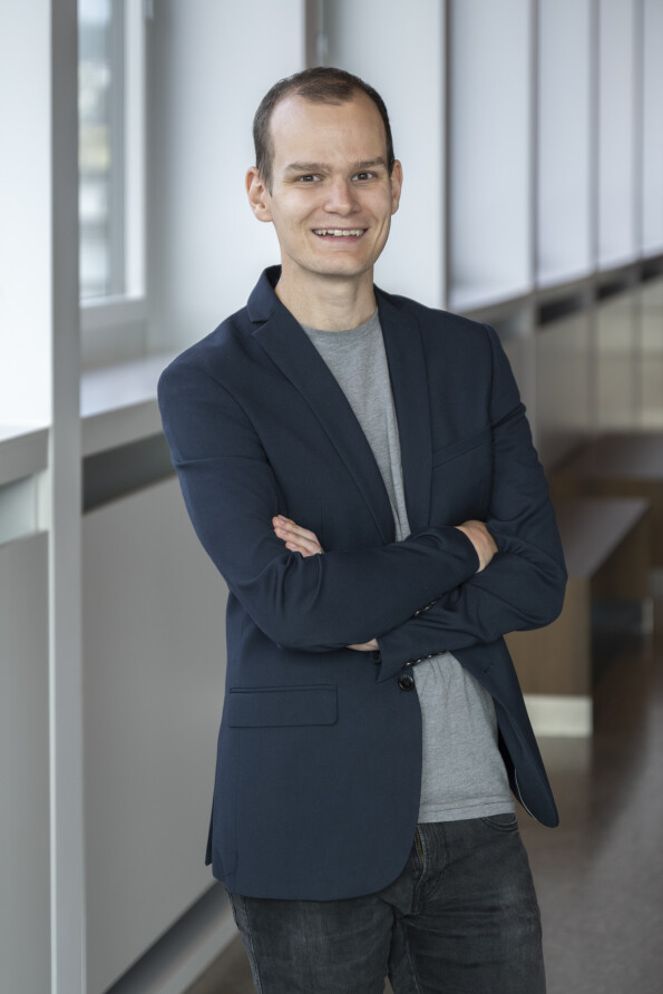 Ein junger Mann mit kurzen Haaren und freundlichem Lächeln steht in einem modernen Büroflur. Er trägt ein dunkelblaues Sakko, ein graues T-Shirt und dunkle Jeans.